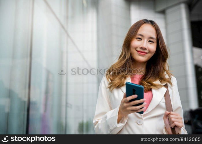 Asian businesswoman texting smartphone commuting work she walking near her office building, Happy business woman smiling using mobile phone outdoor walking on city street urban looking to camera