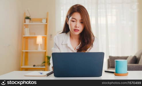 Asian businesswoman using laptop talk to colleagues about plan in video call while smart working from home at living room. Self-isolation, social distancing, quarantine for coronavirus prevention.