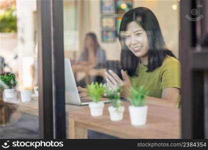 Asian businesswoman working from home with mobile phone and technology laptop computer in happiness feeling at desk beside the glass in modern coffee shop or office,Business owner and entrepreneur concept