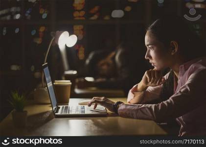 Asian businesswoman working hard late with her technology laptop in office, customer service and call center department, hard worker and overtime, her colleagues sleepy, low light environment,