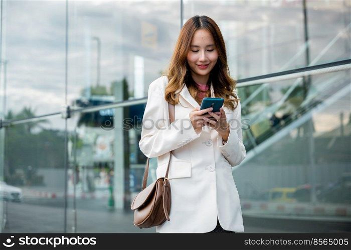 Asian businesswoman working with mobile phone standing against street front building near office, Portrait successful business woman smiling holding smartphone use application chat online in morning