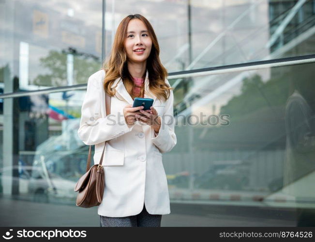 Asian businesswoman working with mobile phone standing against street front building near office, Portrait successful business woman smiling holding smartphone use application chat online in morning