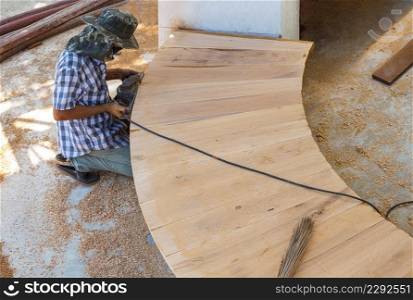Asian carpenter using electric planer to shaving edge of curved wooden bench at workplace in public park area