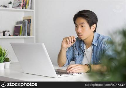 Asian Casual Businessman or Freelancer in Denim or Jeans Shirt Hold Eyeglasses and Working with Laptop in Home Office. Serious casual businessman or Freelancer working with technology