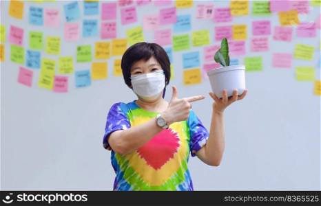 Asian cheerful woman in rainbow t-shirt with protective mask showing her little houseplant with blurred background of sticky notes on gray cement wall at home