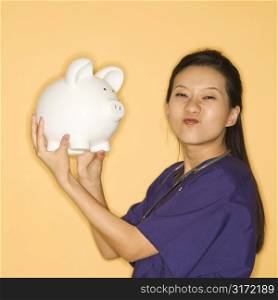 Asian Chinese mid-adult female doctor holding piggy bank against yellow background scrunching nose and looking at viewer.