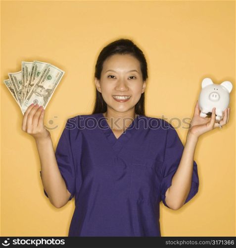 Asian Chinese mid-adult female doctor holding piggy bank in one hand and cash in the other against yellow background smiling and looking at viewer.