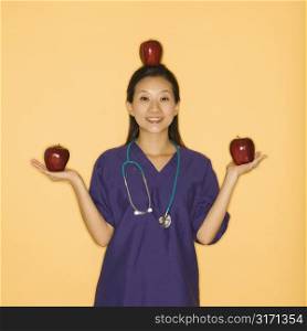 Asian Chinese mid-adult female doctor holding two red apples in hands and balancing one on head against yellow background smiling and looking at viewer.