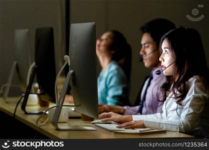 Asian confidence operator woman agent with headsets working in a call center at night Environment with her colleague team as customer service and technical support. Using as Late night Hard working 24 Hr. 7 Days concept.
