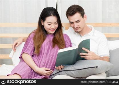 Asian couple family young mother and husband reading book at home, woman and man is pregnant lying on sofa and relax.