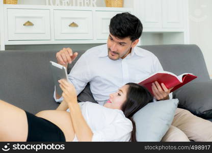 Asian couple family young mother and husband using a tablet at home, woman and man is pregnant lying on sofa looking tablet read book.