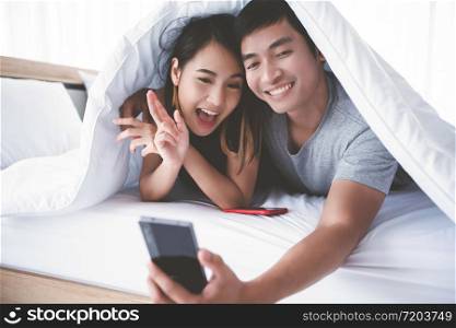 Asian couple lovers lying on their play mobile phones and selfie on bed at home