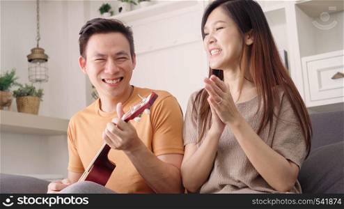 Asian couple playing guitar and singing together in living room at home, sweet couple enjoy love moment while lying on the sofa when relax at home. Lifestyle couple relax at home concept.