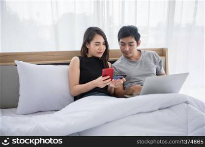 Asian Couple shopping online and paying with credit card on laptop and mobile phone at home,Happy couple at home surfing the net in bed