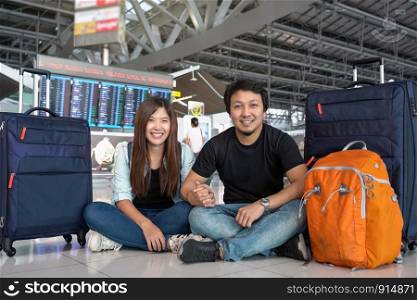 Asian couple traveler sitting with luggage over the flight board for check-in at the flight information screen in modern an airport, travel and transportation concept