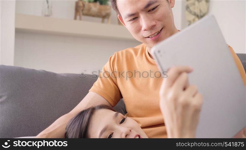 Asian couple using tablet for online shopping in internet in living room at home, sweet couple enjoy love moment while lying on the sofa when relax at home. Lifestyle couple relax at home concept.