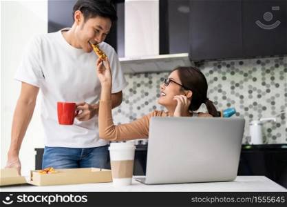 Asian couple work from home in the kitchen and eating delivery pizza take away food and take out coffee while city lockdown from coronavirus covid-19 pandemic. New Normal lifstyle working from home.