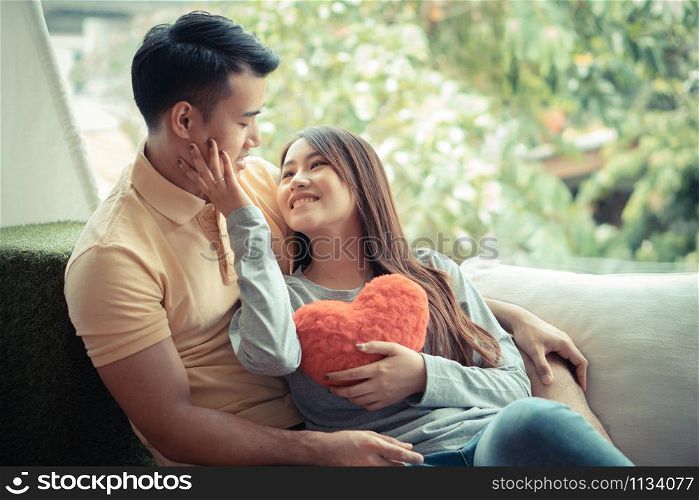 Asian Couples Sitting on the sofa In which women Holding a red heart And smiling happily. Concept of expressions of love and warmth for lovers, Valentine day.