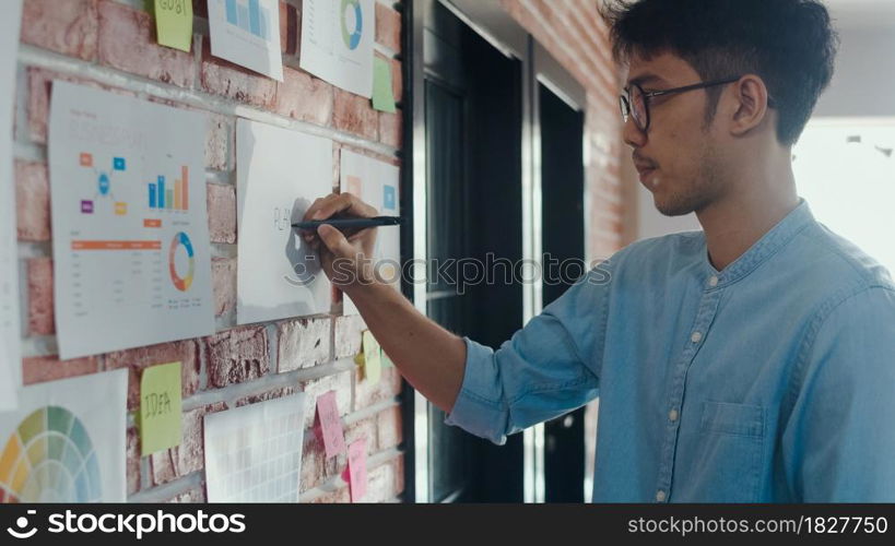 Asian creative man drawing work plan on paper board. Young professional business male think and write information reminder on paper on brick, business situation, startup in Loft office concept.