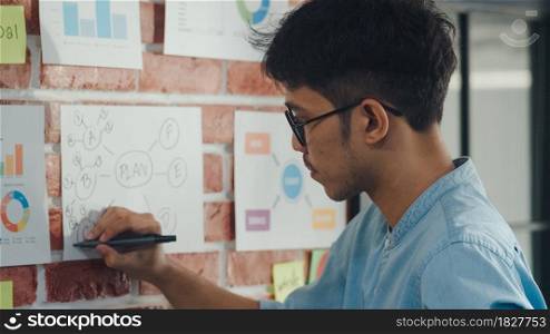 Asian creative man drawing work plan on paper board. Young professional business male think and write information reminder on paper on brick, business situation, startup in Loft office concept.