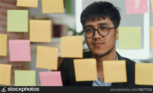 Asian creative man stick a sticky note on glass board. Young professional business male write information, strategy, reminder on paper, business situation, startup in Loft office concept.