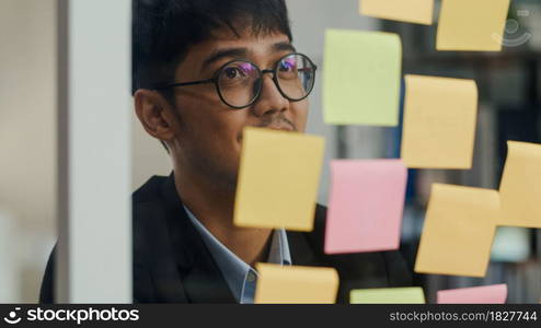 Asian creative man stick a sticky note on glass board. Young professional business male write information, strategy, reminder on paper, business situation, startup in Loft office concept.