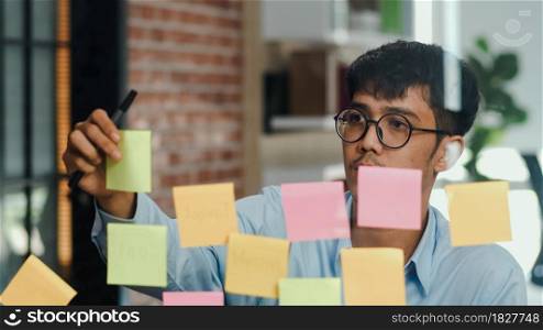 Asian creative man stick a sticky note on glass board. Young professional business male write information, strategy, reminder on paper, business situation, startup in Loft office concept.