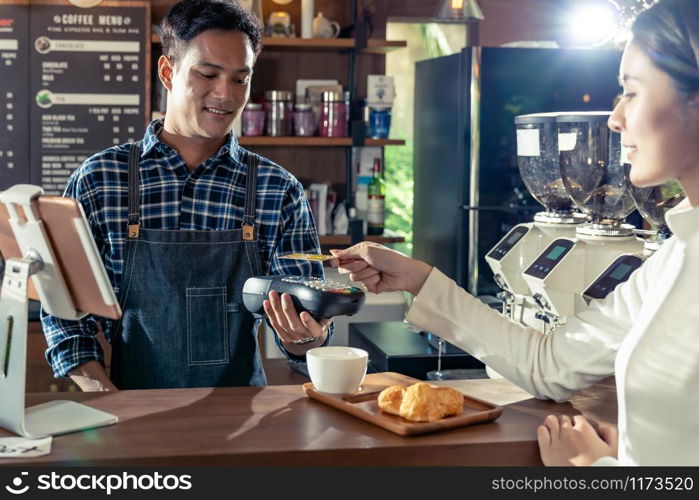 Asian customer using her credit card with contactless nfs technology to pay a barista for her coffee purchase at a cafe bar.