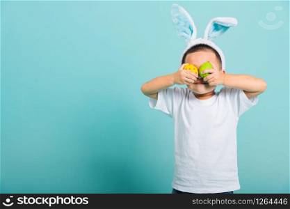 Asian cute little child boy smile beaming wearing bunny ears and a white T-shirt, standing to holds colored easter eggs instead of eyes on blue background with copy space for text