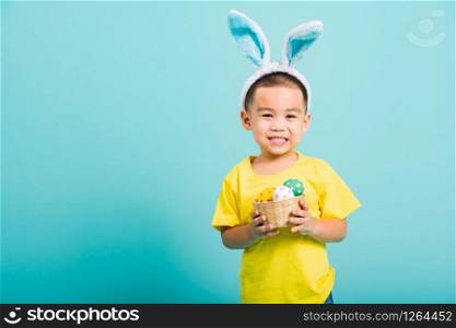 Asian cute little child boy smile beaming wearing bunny ears and a yellow T-shirt, standing to hold a basket with full Easter eggs on blue background with copy space