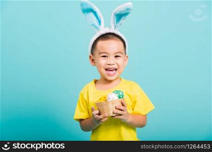 Asian cute little child boy smile beaming wearing bunny ears and a yellow T-shirt, standing to hold a basket with full Easter eggs on blue background with copy space