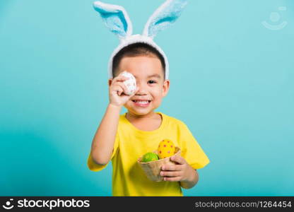 Asian cute little child boy smile beaming wearing bunny ears and a yellow T-shirt, standing to holds colored easter eggs instead of eyes on blue background with copy space for text