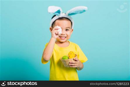 Asian cute little child boy smile beaming wearing bunny ears and a yellow T-shirt, standing to holds colored easter eggs instead of eyes on blue background with copy space for text