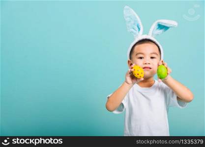 Asian cute little child boy smile beaming wearing bunny ears and a white T-shirt, standing using hand holding and show colored easter eggs on blue background with copy space