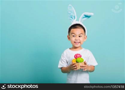 Asian cute little child boy smile beaming wearing bunny ears and a white T-shirt, standing to hold a basket with full Easter eggs on blue background with copy space