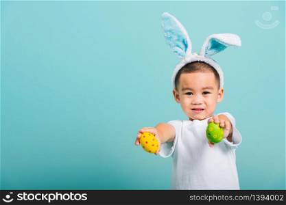 Asian cute little child boy smile beaming wearing bunny ears and a white T-shirt, standing using hand holding and show colored easter eggs on blue background with copy space