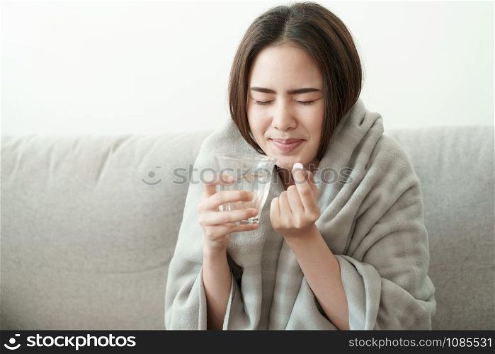 Asian Cute of girl having headache and high temperature from illness holding pill of medicine and water sitting on sofa at home, Health and illness concepts