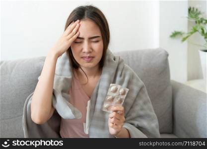 Asian Cute of girl having headache and high temperature from illness holding pill of medicine sitting on sofa at home, Health and illness concepts