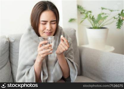 Asian Cute of girl having headache and high temperature from illness holding pill of medicine and water sitting on sofa at home, She dislike to have a medicine, Health and illness concepts