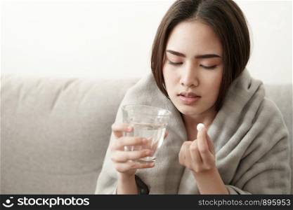 Asian Cute of girl having headache and high temperature from illness holding pill of medicine and water sitting on sofa at home, Health and illness concepts