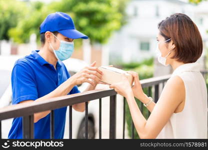 Asian deliver man in blue shirt handling package and give to young woman costumer at house. They wear face mask to reduce coronavirus COVID-19 spreading. Package shopping delivery concept.