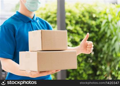 Asian delivery express courier young man use giving boxes to woman customer he wearing protective face mask at front home and thumb up finger, under curfew quarantine pandemic coronavirus COVID-19
