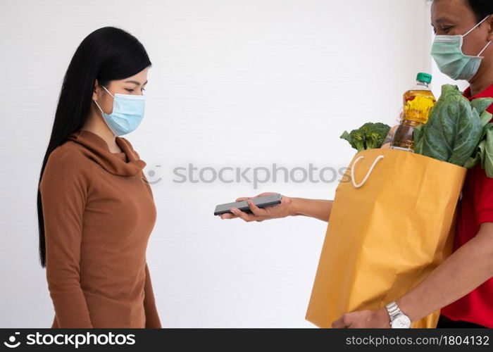 Asian delivery man holding holding a bag of fresh food for giving to customers and holding smartphone for Receive payments at home. Concept of express grocery service and new lifestyle