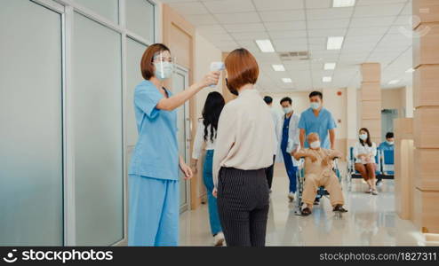 Asian doctor walk through hospital hallway with helper staff push patient wheelchair, operator check fever by digital thermometer visitor at information counter for scan and protect from coronavirus.