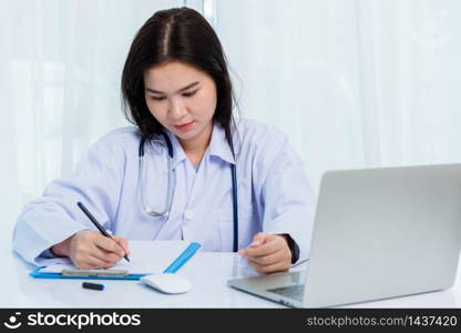 Asian doctor young beautiful woman smiling using working with a laptop computer and her writing something on paperwork or clipboard white paper at hospital desk office, Healthcare medical concept