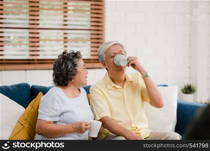 Asian elderly couple drinking warm coffee and talking together in living room at home, couple enjoy love moment while lying on sofa when relaxed at home. Lifestyle senior family at home concept.