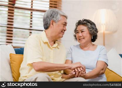 Asian elderly couple holding their hands while taking together in living room, couple feeling happy share and support each other lying on sofa at home. Lifestyle Senior family at home concept.