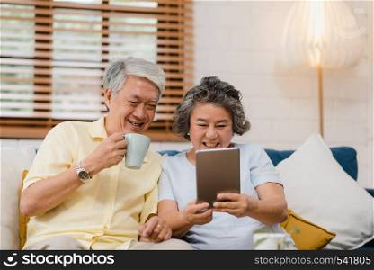 Asian elderly couple using tablet and drinking coffee in living room at home, couple enjoy love moment while lying on sofa when relaxed at home. Enjoying time lifestyle senior family at home concept.
