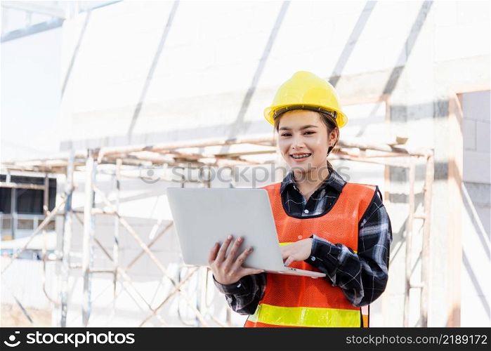 Asian engineer architect worker woman holding laptop inspect and oversee infrastructure progress at construction site, engineering use computer to operate project and control worker to building
