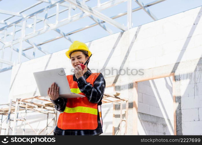 Asian engineer architect worker woman working at build construction site use laptop and talking with radio, engineering hold computer and radio discuss operate and control worker employee to building